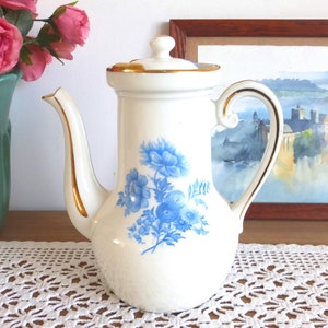 Könnte beinhalten: Weiße Kaffeekanne aus Keramik mit goldener Verzierung und blauem Blumendesign. Die Kanne hat einen Deckel und einen Henkel.