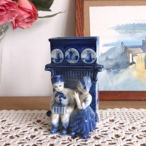 Puede incluir: Jarrón de cerámica azul y blanco con forma de casa holandesa con una figura de niño y niña en la parte delantera. El jarrón tiene tres paneles redondos con imágenes de molinos de viento y barcos.