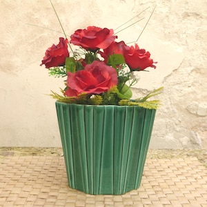 May include: A decorative arrangement of red roses in a green, ribbed ceramic pot. The pot is a trapezoid shape. The roses are surrounded by green foliage and small sprigs. The background is a neutral, textured wall.