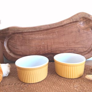 Puede incluir: Dos ramequines de cerámica amarilla con interiores blancos frente a una gran tabla de servir de madera. Los ramequines tienen un diseño acanalado. Flores secas y un tarro decorativo en el fondo.