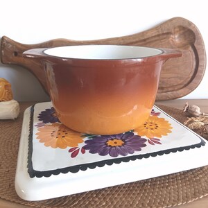 Peut inclure: Une marmite vintage à deux poignées, marron et orange ombré, repose sur un dessous-de-plat carré blanc décoré de motifs floraux colorés. Une planche à découper en bois est à l'arrière-plan.