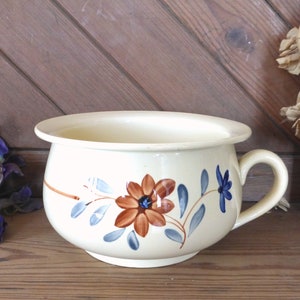 Peut inclure: Un pot de chambre en céramique blanche avec un motif floral bleu et brun. Le pot a une poignée sur le côté et une large ouverture ronde.