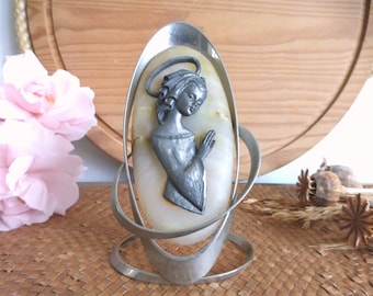 May include: A religious figurine featuring a silver-colored relief of a praying figure with a halo, set against an oval, cream-colored stone. The stone is framed by a silver metal stand with a decorative design. The background includes pink flowers and a woven surface.