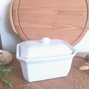 Puede incluir: Una mantequera de cerámica blanca con tapa. El plato tiene forma rectangular con un diseño ligeramente angulado y un pequeño pomo redondo en la tapa. El fondo incluye una tabla de cortar de madera y objetos decorativos.