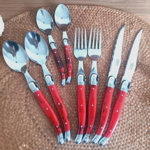 以下が含まれることがあります： スプーン、フォーク、ナイフを含む、赤いハンドルが付いたカトラリーセット。カトラリーはステンレス製で、磨かれた仕上げになっています。