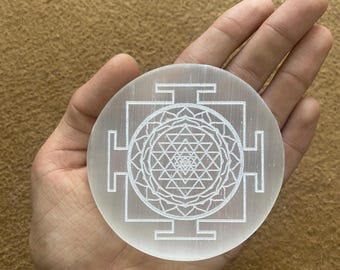 Puede incluir: Un cristal de selenita blanco y redondo con un diseño geométrico detallado grabado en su superficie. El diseño presenta un patrón central similar a un mandala rodeado de formas cuadradas y rectangulares. El cristal se sostiene en una mano abierta.