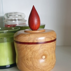 Op de afbeelding: Een handgemaakte houten doos met een druppelvormig deksel. De doos is lichtbruin met een donkerrode band in het midden. Het druppelvormige deksel is dieprood. De doos staat op een witte plank.
