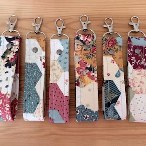 May include: Seven fabric keychains with silver lobster claw clasps. Each keychain has a unique patchwork design with floral, geometric, and plaid patterns in red, blue, yellow, and green. The keychains are displayed on a light brown wooden surface.