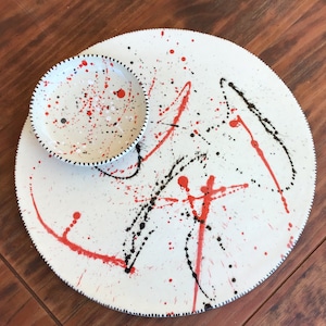 May include: A white ceramic serving dish with a smaller bowl attached. The dish is decorated with red and black splatters of paint.