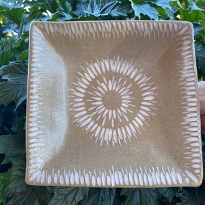 May include: Square ceramic plate with a beige background and a white spiral design in the center. The edges feature a white, repeating pattern. The plate is held by a person's hands, with green foliage in the background.
