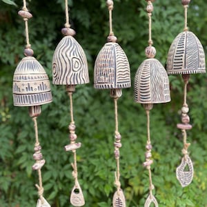 May include: Five handcrafted ceramic wind chimes with bell-shaped tops and intricate carved designs. Each chime hangs from a natural rope, adorned with wooden beads and decorative ceramic pendants. The chimes are a mix of beige and brown tones, creating a rustic, earthy aesthetic.