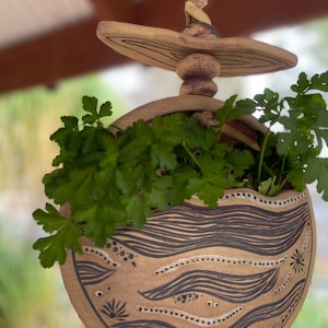 May include: A hanging ceramic planter filled with green parsley. The planter is tan with black wave and dot patterns. It is suspended by a natural fibre rope. The top of the planter has a decorative mushroom-shaped element.