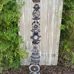 May include: A black and white ceramic garden totem with a variety of geometric shapes and patterns. The totem is made up of several stacked pieces, each with a unique design. The totem is standing on a brown surface.