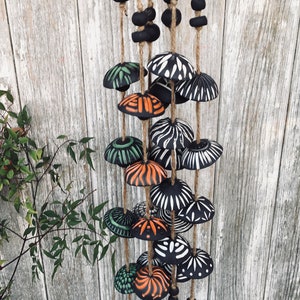 May include: A black and white wind chime with orange, green, and black accents. The chime is made of ceramic and has a variety of geometric patterns. It is hanging from a wooden surface.