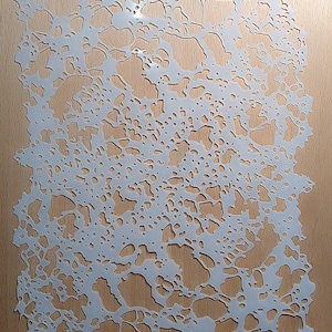 May include: A white, intricate stencil with a network of organic, irregular shapes, resembling a natural pattern. The stencil is placed on a light brown wooden surface, with a small, dark metal fastener at the top.