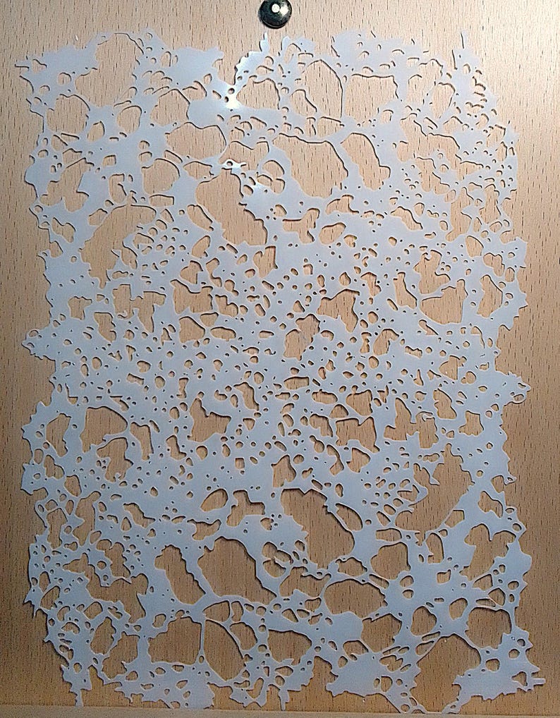May include: A white, intricate stencil with a network of organic, irregular shapes, resembling a natural pattern. The stencil is placed on a light brown wooden surface, with a small, dark metal fastener at the top.