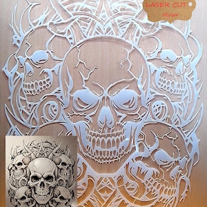 May include: A laser-cut, white mylar skull design with intricate details. The design features multiple skulls and decorative elements. The text "Durable Laser Cut Mylar" is visible. The design is mounted on a light brown surface.
