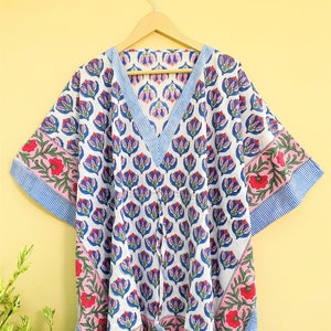 Op de afbeelding: Een witte kaftan met een V-hals, versierd met een bloemmotief van blauwe en roze bloemen. De mouwen en de halslijn zijn afgezet met blauw-witte strepen. Het heeft een taille met trekkoord en hangt aan een houten hanger.