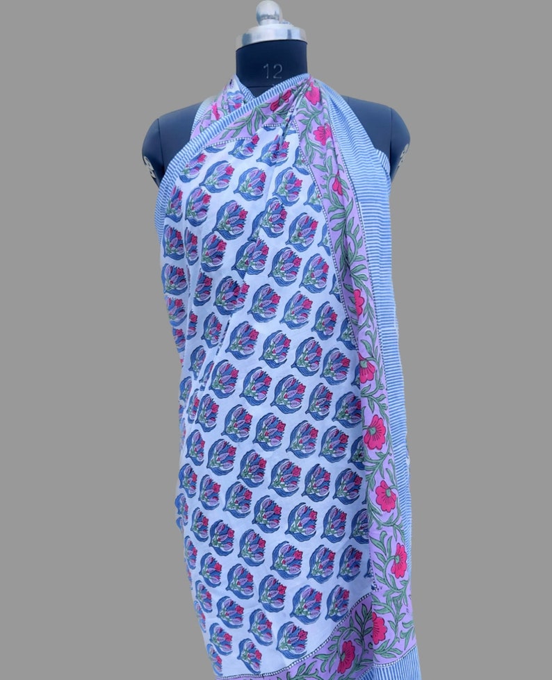 Puede incluir: Un pareo blanco con un estampado floral repetido en azul, morado y rosa. El pareo est&aacute; drapeado sobre un maniqu&iacute;. El borde presenta un dise&ntilde;o a rayas en azul y blanco, con detalles florales en rosa y verde.