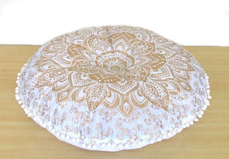 May include: Round floor cushion with a gold and white mandala design. The cushion has a buttoned center and is trimmed with white pom-poms. The cushion is approximately 30 inches in diameter.