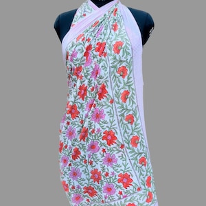 Puede incluir: Un pareo blanco con un estampado floral de flores rojas y rosas y hojas verdes. El pareo tiene un borde a rayas rosa claro. La tela est&aacute; drapeada sobre un maniqu&iacute; negro.