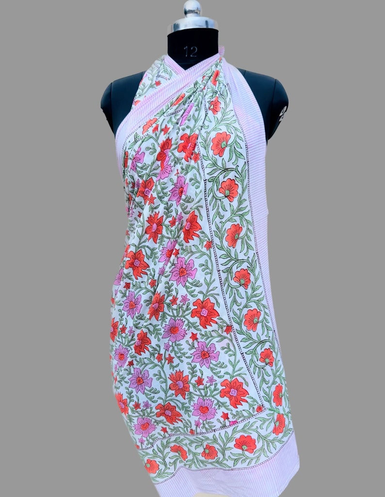 Puede incluir: Un pareo blanco con un estampado floral de flores rojas y rosas y hojas verdes. El pareo tiene un borde a rayas rosa claro. La tela est&aacute; drapeada sobre un maniqu&iacute; negro.