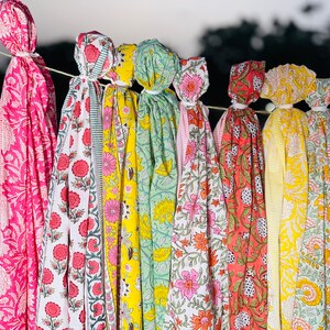 May include: A collection of colourful fabric scarves with floral patterns, hanging on a line. The scarves are in shades of pink, yellow, green, and orange.