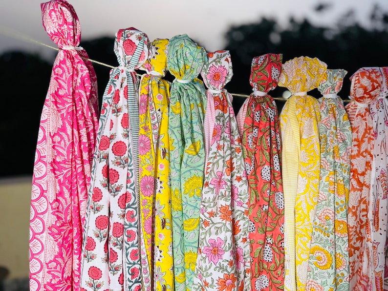 May include: A collection of colourful fabric scarves with floral patterns, hanging on a line. The scarves are in shades of pink, yellow, green, and orange.