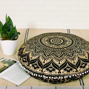 May include: A round floor cushion with a black and gold mandala design. The cushion is trimmed with white pom-poms. A potted snake plant and an open book are also in the image.