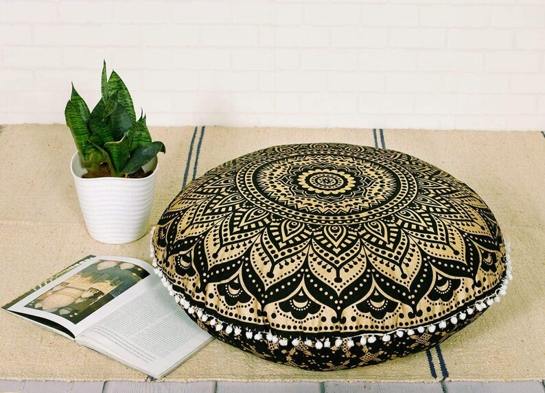 May include: A round floor cushion with a black and gold mandala design. The cushion is trimmed with white pom-poms. A potted snake plant and an open book are also in the image.