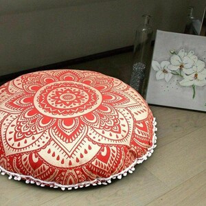 May include: A round floor cushion with a red and cream mandala design. The cushion is trimmed with white pom-poms. The cushion is a decorative item for a living room or bedroom.