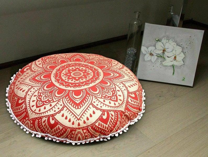 May include: A round floor cushion with a red and cream mandala design. The cushion is trimmed with white pom-poms. The cushion is a decorative item for a living room or bedroom.