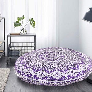 May include: A large, round floor cushion with a detailed purple and white mandala pattern. The cushion is approximately 36 inches in diameter and features a decorative pom-pom trim. It is placed on a wooden floor in a room with a minimalist aesthetic.