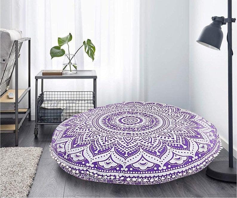 May include: A large, round floor cushion with a detailed purple and white mandala pattern. The cushion is approximately 36 inches in diameter and features a decorative pom-pom trim. It is placed on a wooden floor in a room with a minimalist aesthetic.
