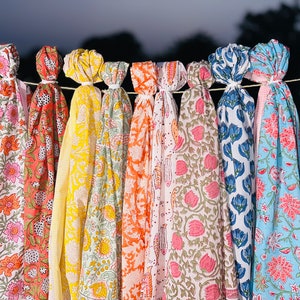May include: A collection of colourful fabric scarves with floral patterns hanging on a clothesline. The scarves are in shades of green, pink, orange, yellow, blue, and white.