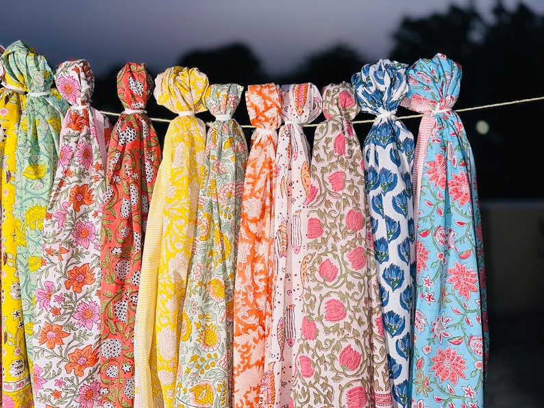 May include: A collection of colourful fabric scarves with floral patterns hanging on a clothesline. The scarves are in shades of green, pink, orange, yellow, blue, and white.