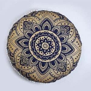 May include: Round decorative cushion with a gold and navy blue mandala design. The cushion has a white pom-pom trim around the edge. The cushion is a decorative item.