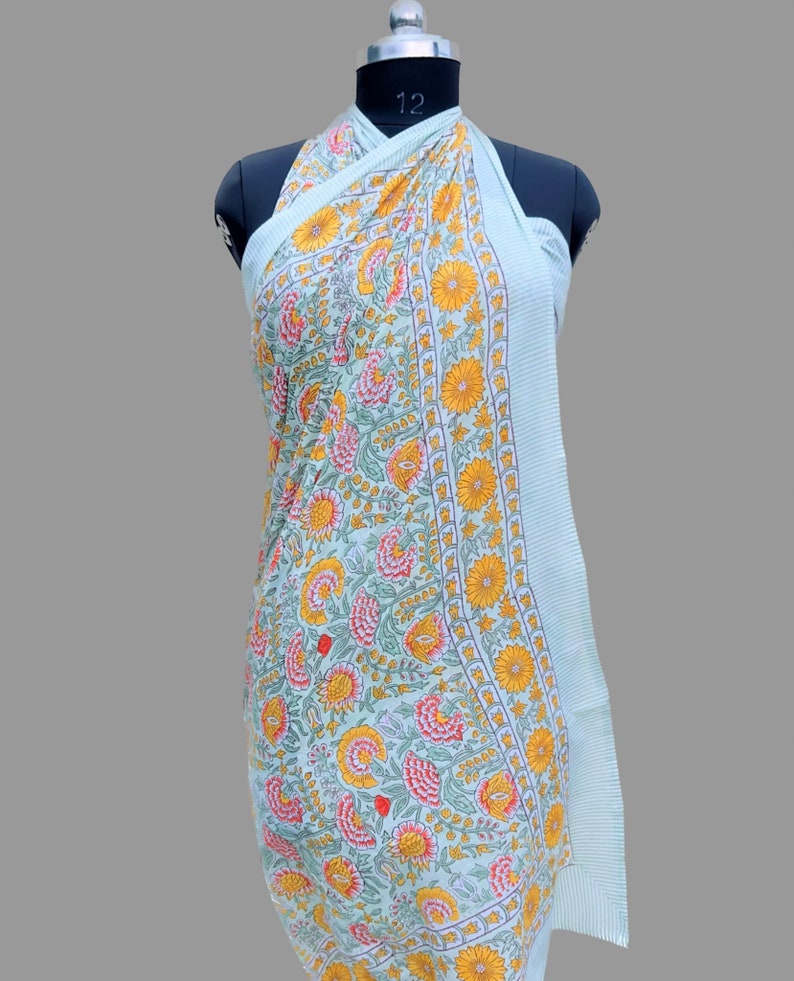 Puede incluir: Un pareo verde claro con un estampado floral de flores amarillas y rosas y hojas verdes. El pareo est&aacute; drapeado sobre un maniqu&iacute; y tiene un borde a rayas.
