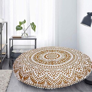 May include: Round floor cushion with a white and gold mandala design. The cushion has a decorative pattern and is trimmed with white pom-poms. The cushion is placed on a wooden floor.