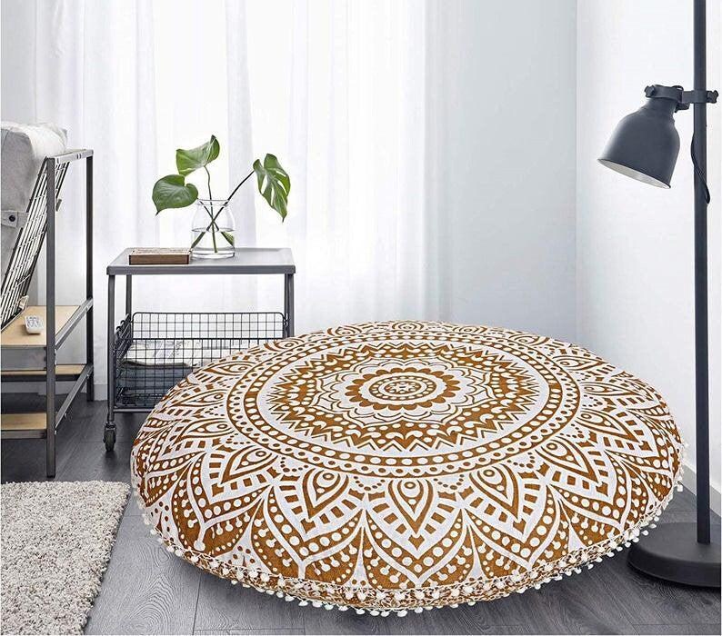 May include: Round floor cushion with a white and gold mandala design. The cushion has a decorative pattern and is trimmed with white pom-poms. The cushion is placed on a wooden floor.