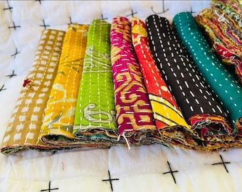 May include: A stack of eight colorful kantha fabric scraps, each with a different pattern and stitching. The fabrics are folded and arranged in a row, with the most colorful fabric at the top.