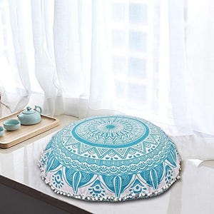 May include: A round floor cushion with a turquoise and white mandala design. The cushion has a white trim with small white pom-poms. The cushion is placed near a window with white curtains.