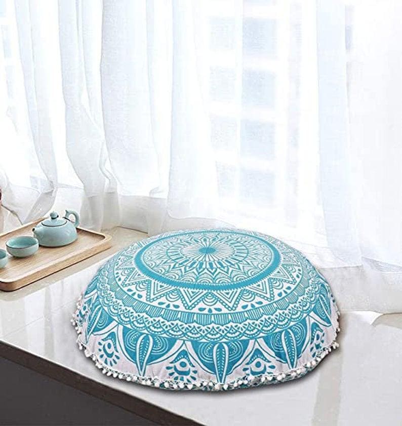 May include: A round floor cushion with a turquoise and white mandala design. The cushion has a white trim with small white pom-poms. The cushion is placed near a window with white curtains.