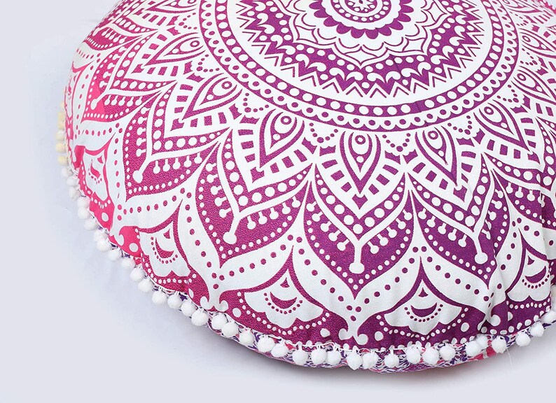 May include: Round floor cushion with a mandala design in shades of purple and white. The cushion has a gradient effect, transitioning from pink to purple. White pom-poms trim the edge. A decorative accent for home decor.
