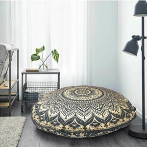 May include: A large, round floor cushion with a black and gold mandala design. The cushion is placed on a hardwood floor in a room with a neutral color scheme. The cushion is approximately 36 inches in diameter.