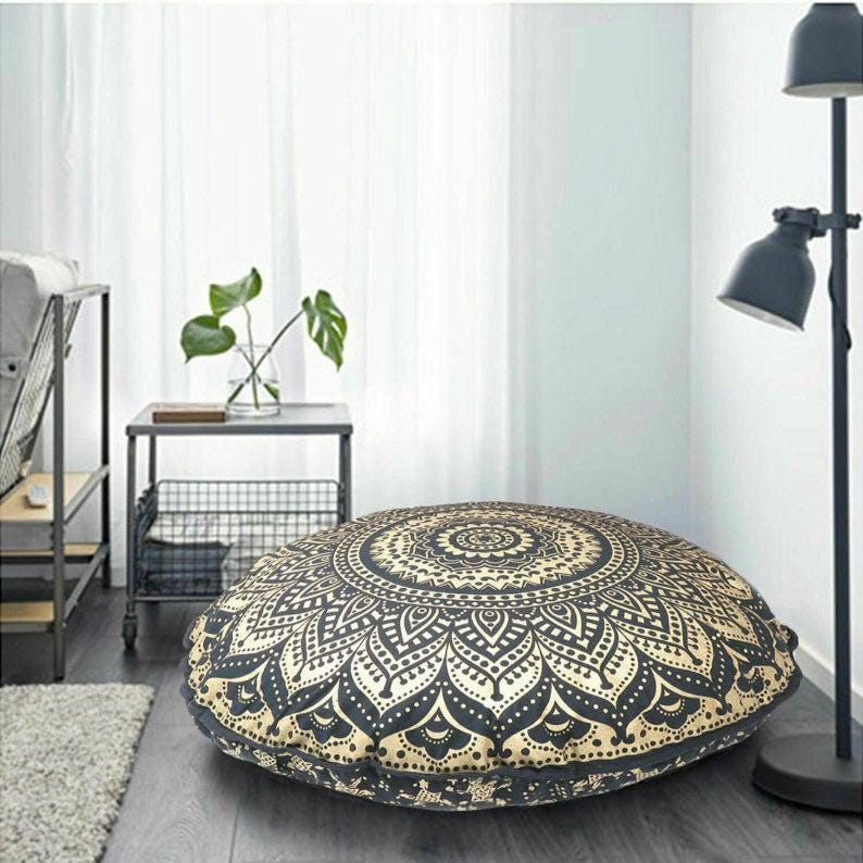 May include: A large, round floor cushion with a black and gold mandala design. The cushion is placed on a hardwood floor in a room with a neutral color scheme. The cushion is approximately 36 inches in diameter.