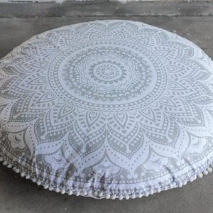 May include: Round floor cushion with a gray and white mandala pattern. The cushion has a white pom-pom trim around the edge. The cushion is on a gray surface.