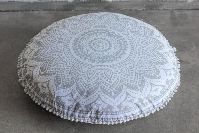 May include: Round floor cushion with a gray and white mandala pattern. The cushion has a white pom-pom trim around the edge. The cushion is on a gray surface.
