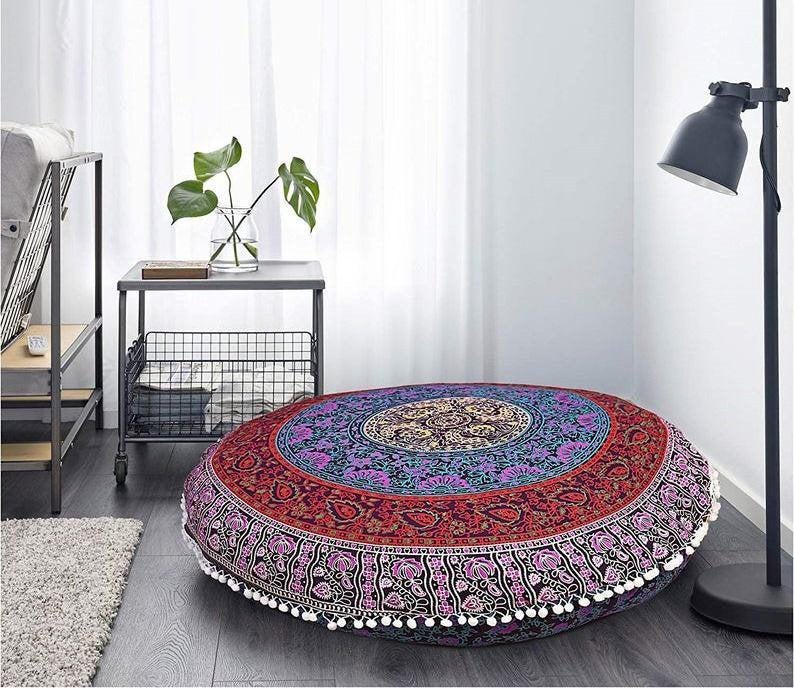 May include: A large, round floor cushion with a vibrant mandala design. The cushion features a central medallion in gold, surrounded by concentric rings of red, purple, and blue. White pom-poms trim the edge. This decorative cushion is perfect for a living room or meditation space.