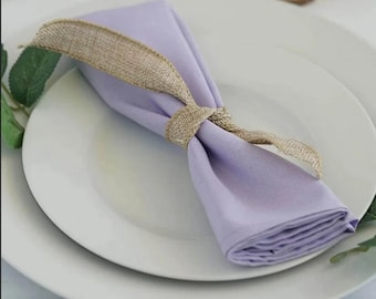 May include: A folded lavender napkin tied with a tan burlap ribbon, presented on a white plate. The napkin is neatly folded and the ribbon is tied in a decorative knot. The setting suggests a formal dining occasion.